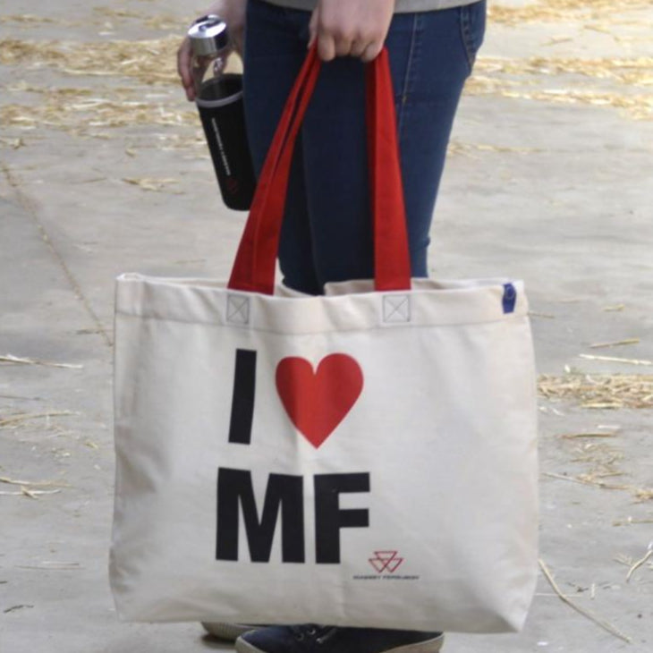 Person holding an AGCO Massey Ferguson shopping bag (Product Code: X993342304000) made of beige cotton with red handles and the text "I ♥ MF" printed on it, while carrying a black drink container.