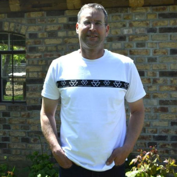A man wearing an AGCO Massey Ferguson White Crew Neck T-Shirt (Product Code: X993412302000) stands outside in front of a brick building with a tiled roof.