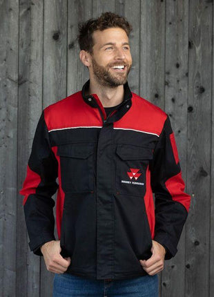 A man with short hair and a beard, adorned in an AGCO Massey Ferguson Black And Red Work Jacket (X993452102), stands in front of a wooden wall. Smiling warmly, his hands are comfortably tucked into his pockets.