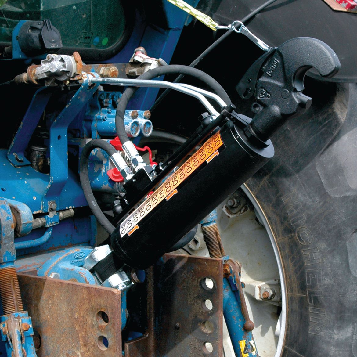 Close-up of a Sparex Hydraulic Top Link (Cat.2/3) Knuckle and Q.R CBM Hook, with a 90mm cylinder bore and minimum length of 690mm, connected to industrial machinery. A visible gauge and several connected hoses are featured. The machinery is painted blue, with a hose set in place, and the background shows part of a black tire. Sparex Part No.S.399732