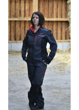 A person wearing the AGCO Massey Ferguson Women's Work Jacket (X993532214), featuring adjustable cuffs and reflective stripes, stands on a concrete floor in a barn with hay scattered in the background.