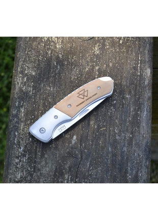 An AGCO Massey Ferguson folding pocket knife, model X993562301000, with a stainless steel blade and a metallic brown handle showcasing a geometric design, placed on a wooden surface.