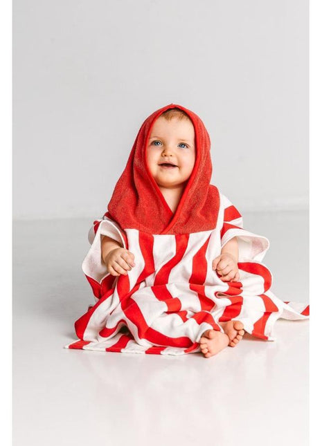 A baby sits on the floor wearing a Massey Ferguson Poncho for Toddlers by AGCO, identified as X993602305000. The red and white striped poncho, made of soft terry cloth, has a red hood that covers the baby's head. The baby appears to be smiling.