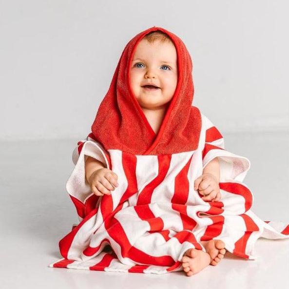 A baby sits on the floor wearing a Massey Ferguson Poncho for Toddlers by AGCO, identified as X993602305000. The red and white striped poncho, made of soft terry cloth, has a red hood that covers the baby's head. The baby appears to be smiling.