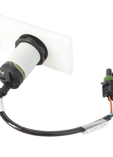 A white cylindrical sensor with a black connector is attached to a multi-colored wire harness and connected to a white mounting plate. This product is the AGCO Switch - Acw6174940 by AGCO. Further product description information is currently not available.