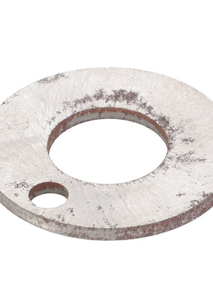 The AGCO | Shim - F181108080013 is a flat, round, metal washer featuring a larger central hole and a smaller secondary hole on one side, both displaying signs of wear and oxidation.