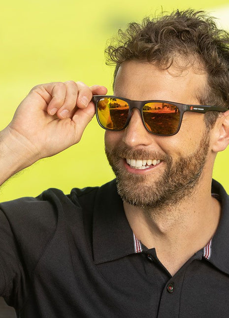 Man with a beard, wearing a black polo shirt and AGCO Massey Ferguson sunglasses (model X993472201000), smiles as he lifts one arm to adjust his UV protection glasses. The background is a bright, blurred green.